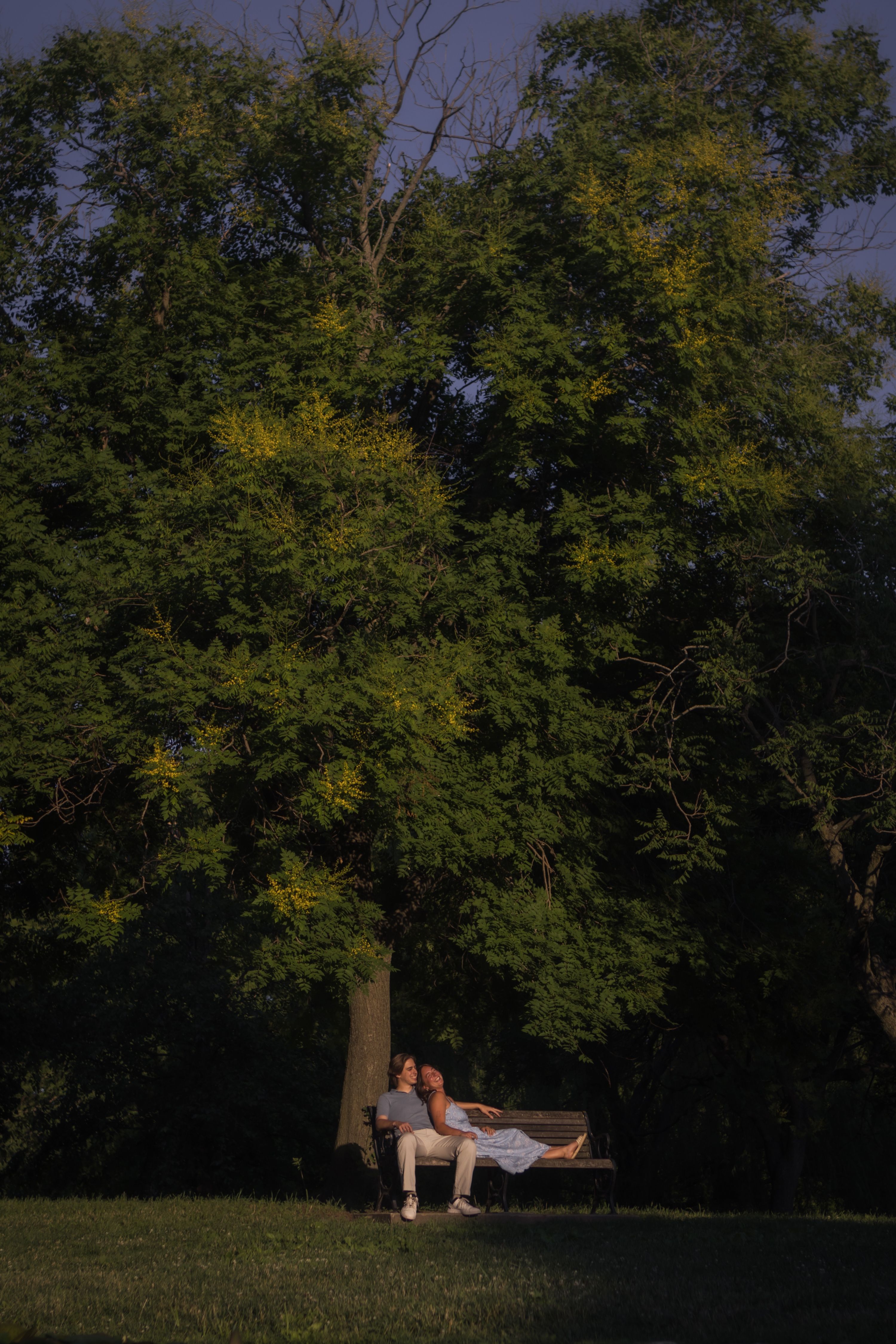 Andrew and Grace on a park bench under a large tree, Grace stretched out along the bench and leaning back against Andrew