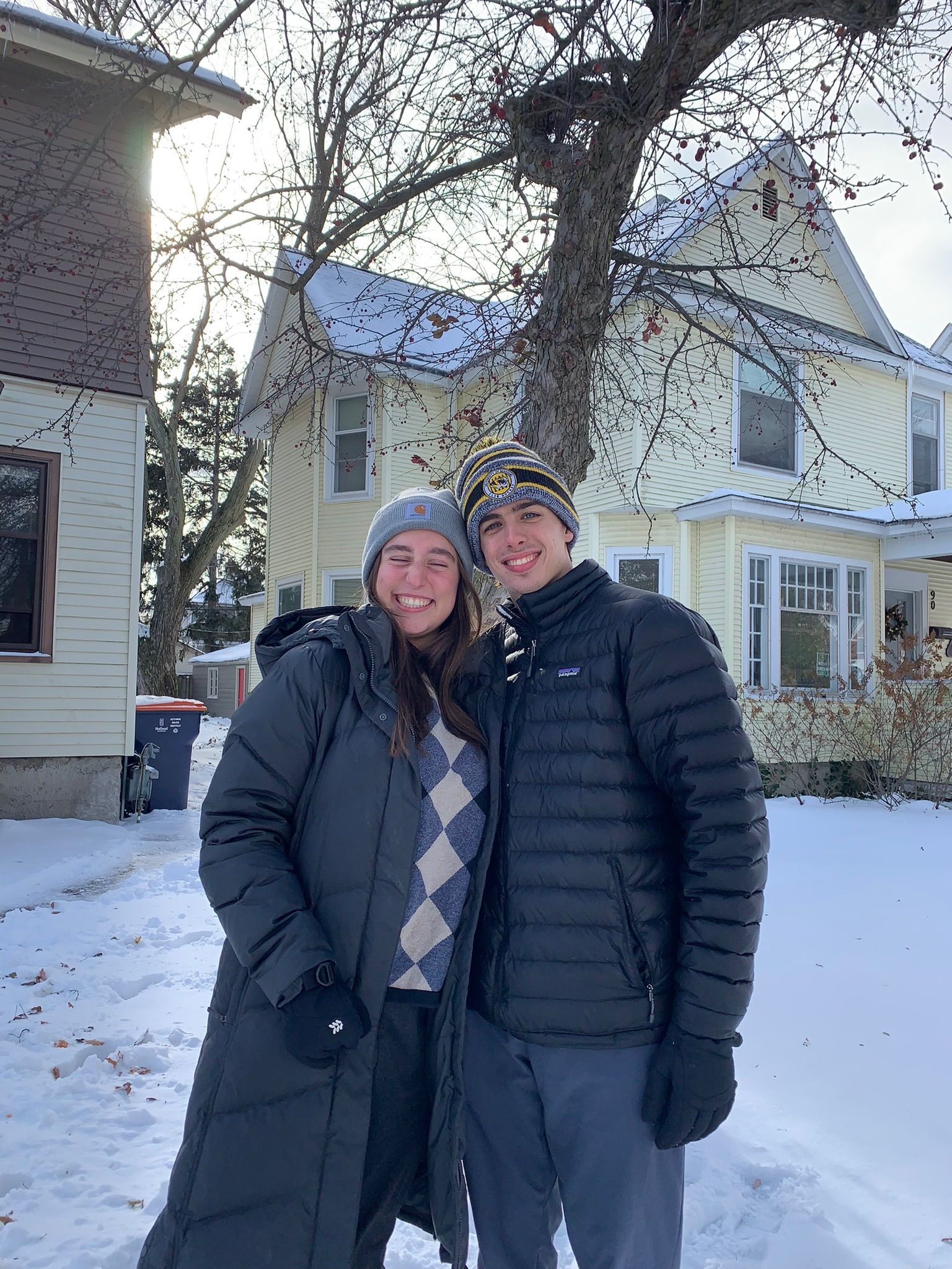 Andrew and Grace in winter coats in front of a snowy yellow house